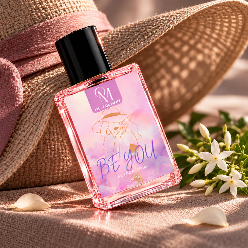 Be You pink perfume bottle angled against a wide-brim woven hat and textured linen, with a jasmine spray in warm morning light.