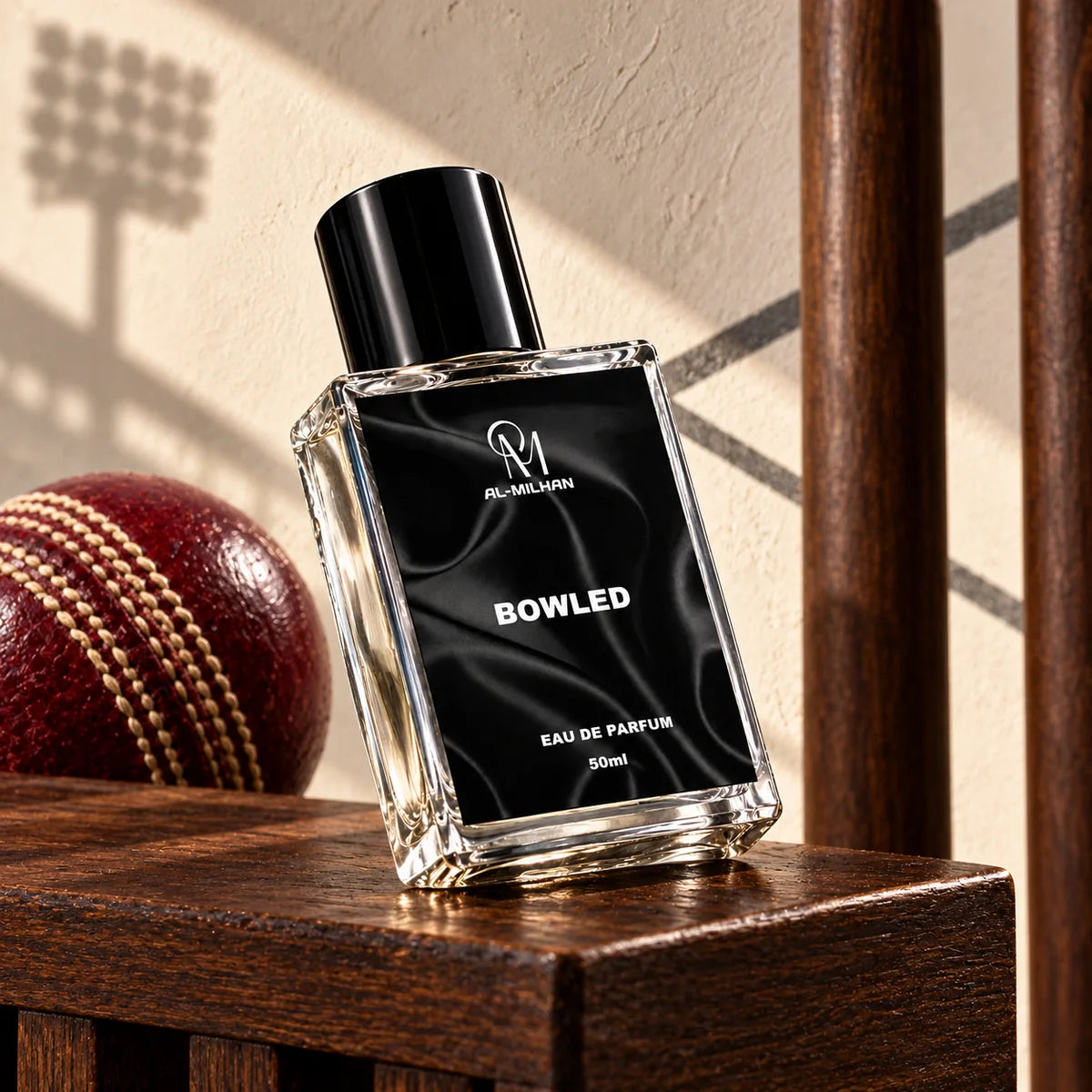 Bowled clear perfume bottle with its black label beside a burgundy stitched cricket ball and wooden wickets against a cream pavilion wall.