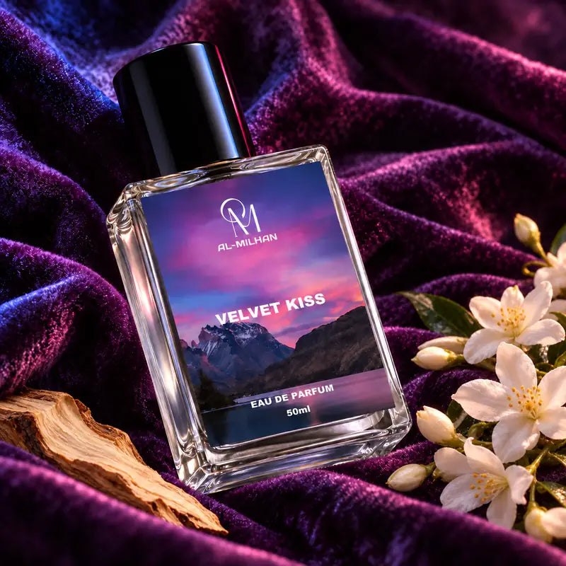Velvet Kiss clear perfume bottle with its sunset-mountain label tilted across violet velvet, with white flowers and a pale wood sliver.